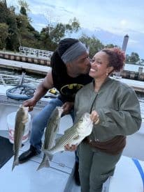 A couple joyfully displaying their catch of striped bass while enjoying a romantic moment on the boat.