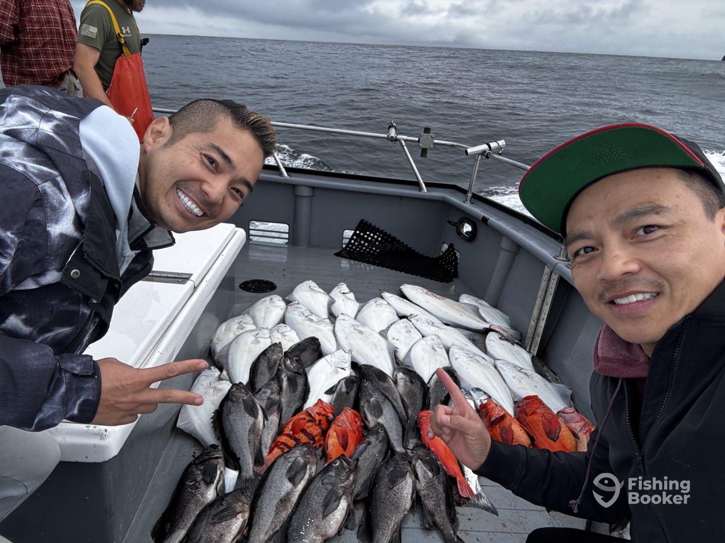 Two anglers smiling and posing with a variety of fish, including Halibut and Rockfish, on a fishing boat in open water.