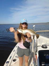 An angler proudly displaying a large Speckled Trout while fishing from a boat in a scenic waterway.