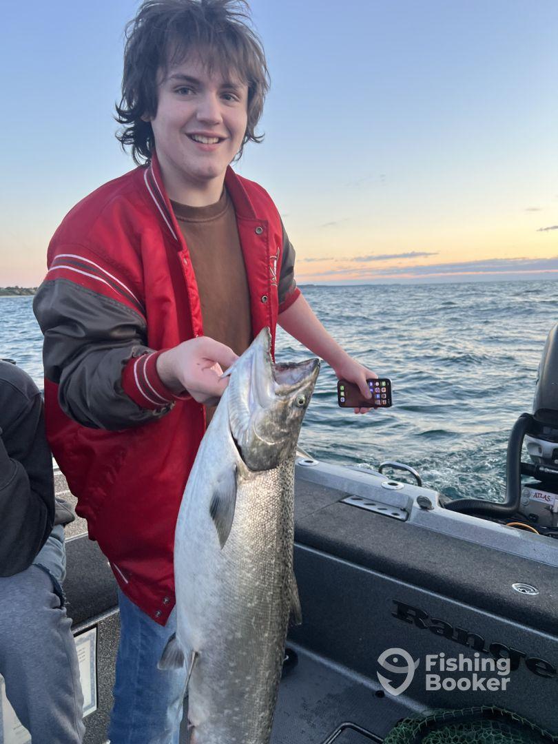 A young angler proudly displaying a large salmon while fishing on a boat during sunset.