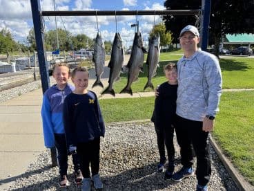 A family poses proudly in front of a catch board displaying several Salmon at a marina, showcasing their successful fishing trip.