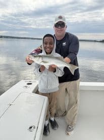 A young boy and an adult proudly displaying a striped bass while fishing on a calm lake, showcasing a memorable catch.