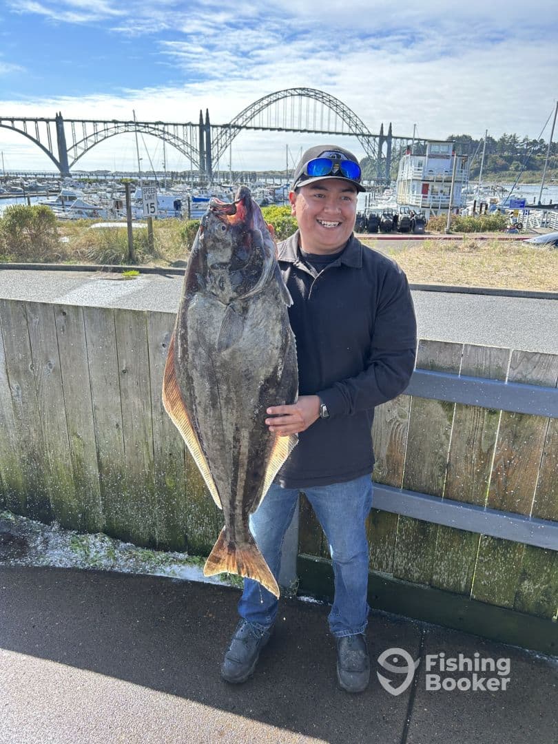 Angler proudly displaying a large Halibut at the dock, with a scenic marina and bridge in the background.