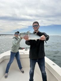 Two anglers on a boat, with one proudly displaying a large fish, likely a Mackerel, while the other is actively fishing in a scenic ocean setting.