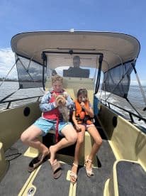 Two children seated inside a boat, enjoying a fishing trip with their small dogs, while an adult operates the helm.