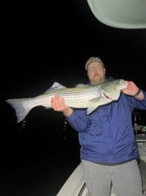 Angler proudly displaying a large Striped Bass while fishing at night, showcasing the excitement of a successful catch.