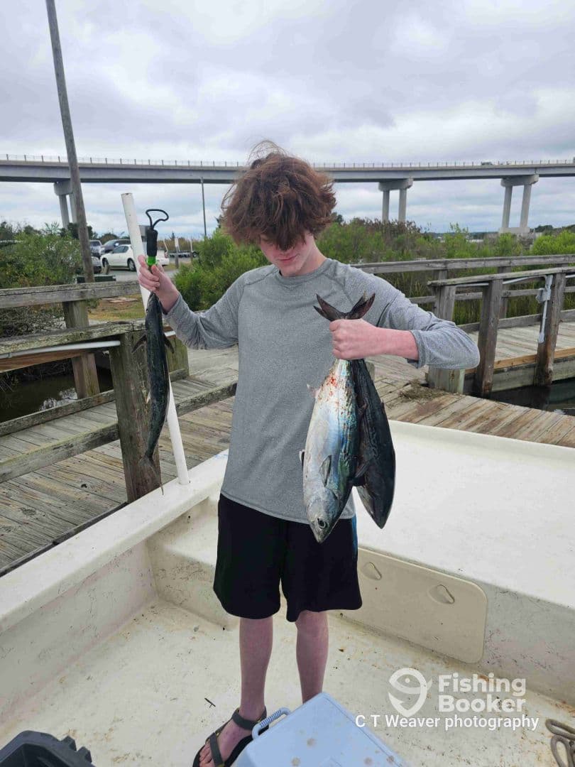 A young angler proudly displaying a large fish while standing on a boat, with a scenic backdrop of a bridge and dock.