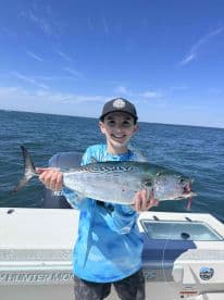 A young angler proudly displaying a large fish while on a fishing trip, with a clear blue sky and ocean in the background.