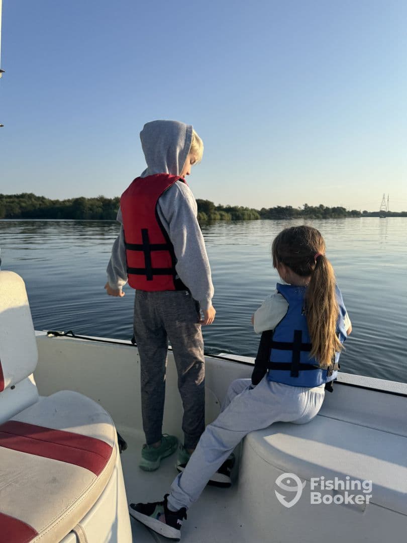 Two children in life jackets enjoying a calm morning on a fishing boat, gazing at the serene water and surrounding nature.
