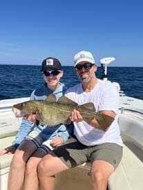 A young angler and his father proudly displaying a large Grouper while fishing on a boat in clear blue waters.