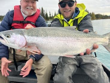 Two anglers proudly displaying a large Rainbow Trout while fishing from a boat in a scenic lake setting.