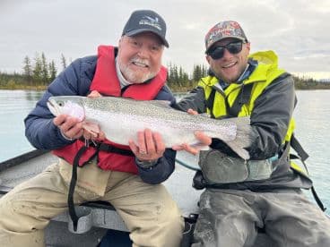 Two anglers proudly displaying a large Rainbow Trout while fishing on a serene lake surrounded by trees.