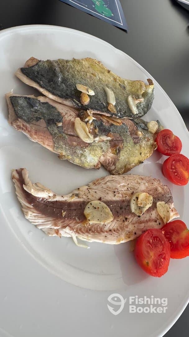 Three fillets of freshly cooked fish garnished with garlic and served with sliced tomatoes, showcasing a delicious meal after a fishing trip.
