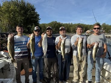 A group of six anglers proudly displaying their catch of Walleye on a sunny day, showcasing a successful fishing trip.