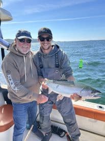 Two anglers proudly displaying a large Salmon aboard a fishing boat, enjoying a sunny day on the water.