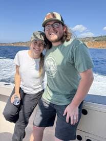 A young couple enjoying a sunny day on a fishing boat, with scenic coastal views in the background.