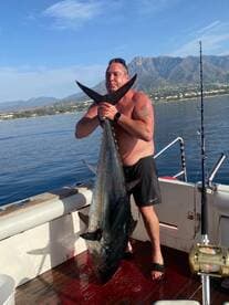 Angler proudly displaying a large fish on a boat, with a scenic backdrop of mountains and calm waters.