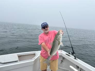 An angler proudly displaying a large Striped Bass while fishing on a foggy day.