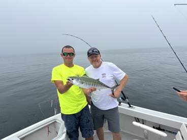 Two anglers proudly displaying a large striped fish aboard a boat in a foggy fishing environment.