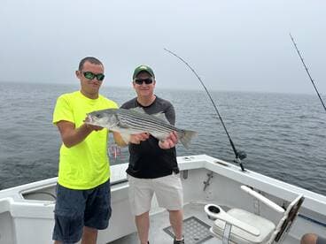 Two anglers proudly displaying a striped bass aboard a fishing boat in foggy conditions.