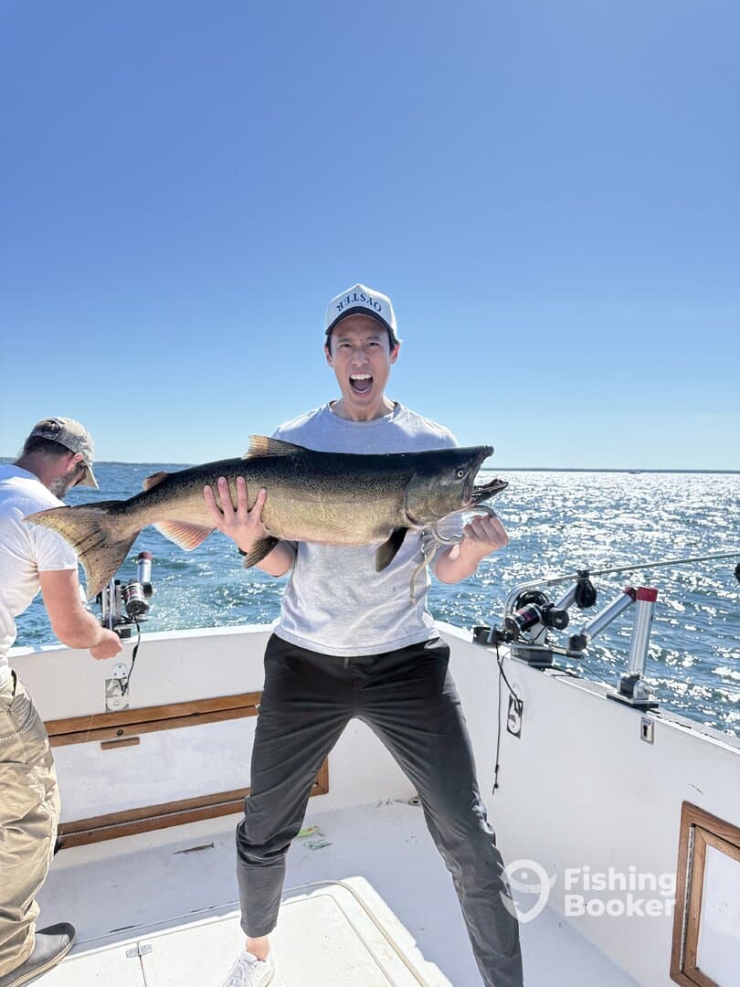 Excited angler holding a large Salmon while fishing on a boat, showcasing a successful catch in bright sunny conditions.