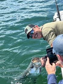 Angler reeling in a large fish while another person captures the moment on a tablet, showcasing an exciting fishing experience on the water.