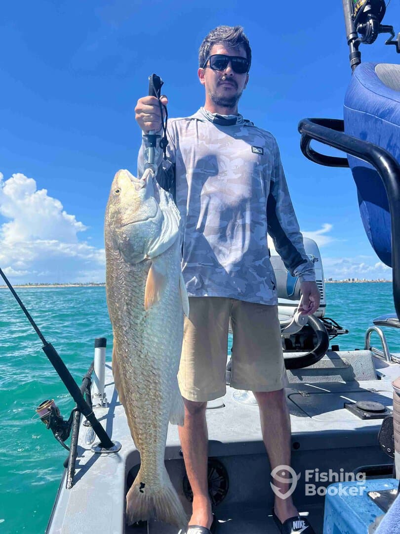Angler proudly displaying a large Redfish while on a fishing boat in clear blue waters.