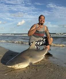 An angler proudly displaying a large shark on the beach, showcasing a successful catch during a coastal fishing trip.
