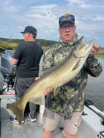 An angler proudly displaying a large trout while fishing on a calm river, with another fisherman in the background.