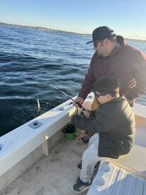 A young angler and an adult are engaged in fishing on a boat, with the child reeling in a catch in a scenic water environment.