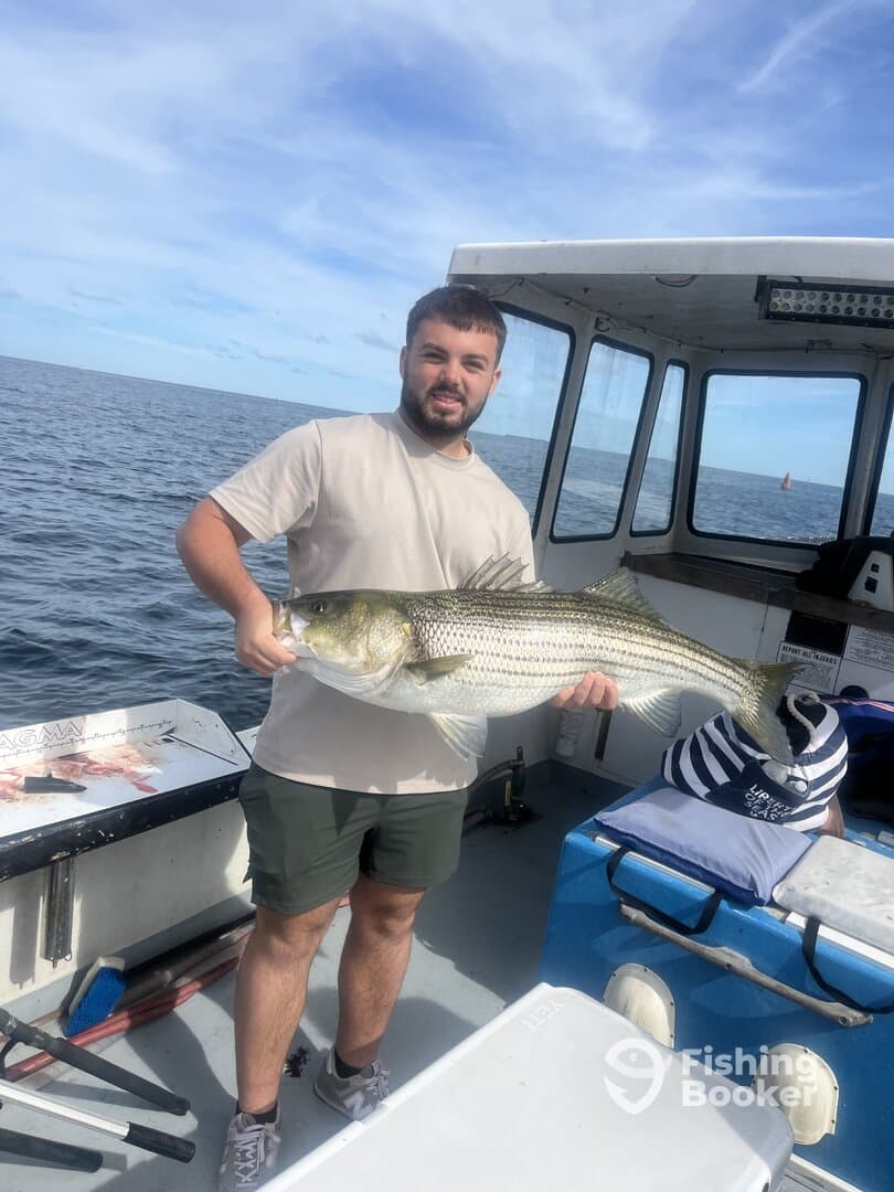 An angler proudly displaying a large Striped Bass aboard a fishing boat, showcasing a successful day on the water.