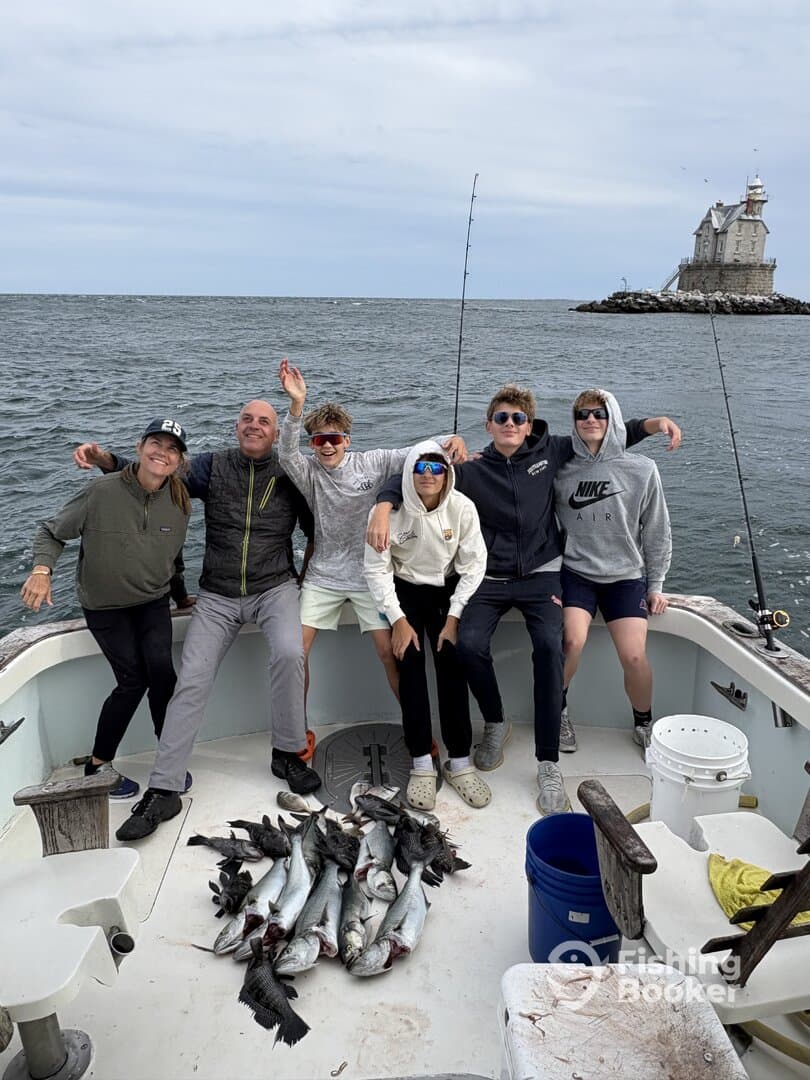 A group of excited anglers, including minors, celebrating their successful catch of multiple fish on a boat, with a scenic lighthouse in the background.
