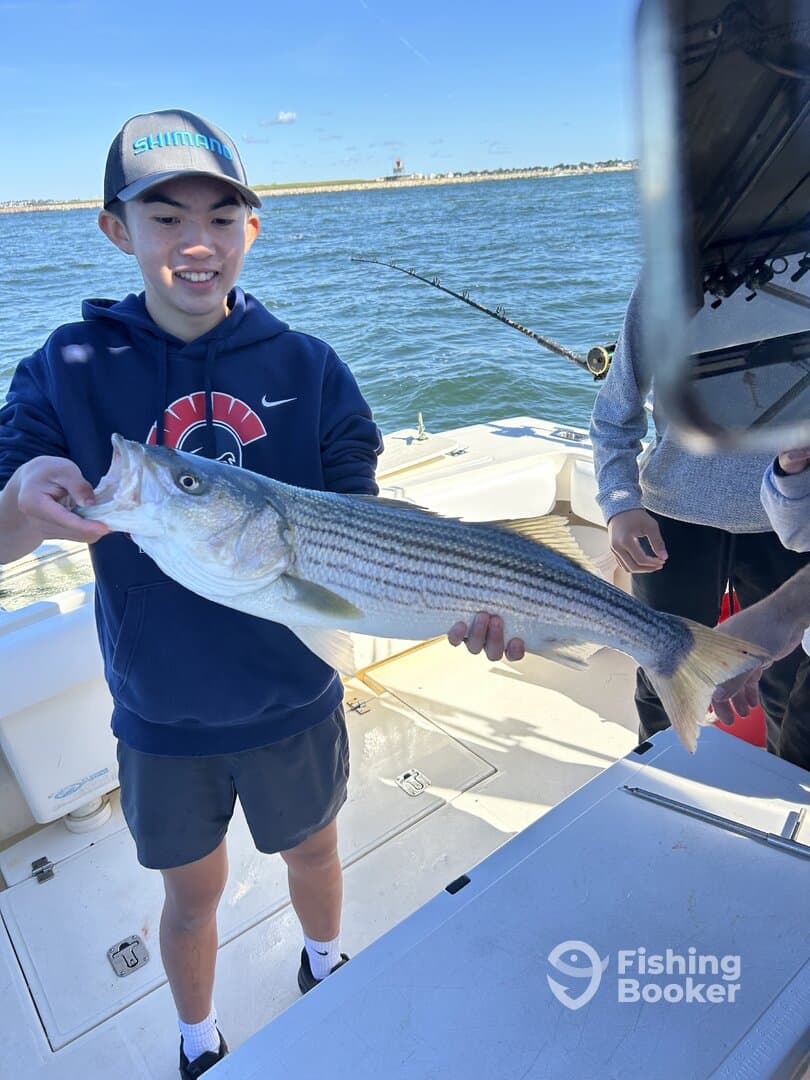 A young angler proudly displaying a large Striped Bass while fishing on a boat, showcasing a successful day on the water.