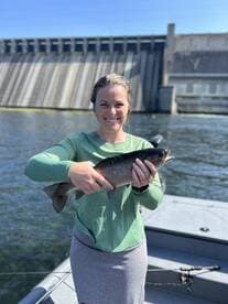 Angler proudly displaying a freshly caught Rainbow Trout while fishing from a boat near a dam.