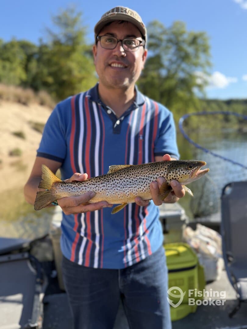 Angler proudly displaying a beautiful Brown Trout while fishing from a boat in a serene lake setting.