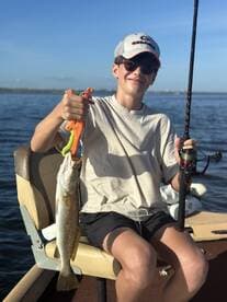 A young angler proudly displaying a Speckled Trout while seated on a boat, enjoying a sunny day on the water.