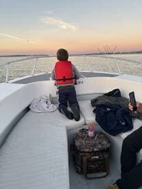 A young boy in a red life jacket gazes out over the water from the interior of a boat, enjoying a serene sunset view.