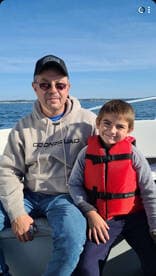 A grandfather and his young grandson enjoying a day on the boat, both smiling while seated in the cabin area, with a scenic ocean view in the background.