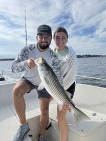 Two anglers proudly displaying a large Striped Bass on a fishing boat, enjoying a successful day on the water.