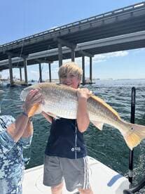A young angler proudly displaying a large Redfish while fishing on a boat under a bridge.