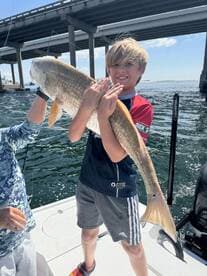 A young angler proudly displaying a large Redfish while fishing under a bridge, showcasing the excitement of a successful catch.