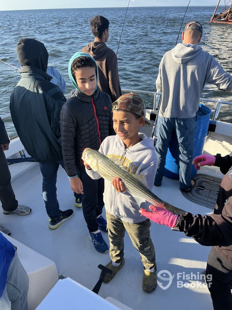 Two young anglers proudly displaying a striped bass while fishing on a boat, surrounded by fellow fishermen.