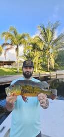 Angler proudly displaying a vibrant Peacock Bass while fishing in a tropical setting, surrounded by palm trees.