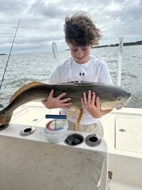 A young angler joyfully displaying a large Redfish on a boat, showcasing a successful fishing trip.