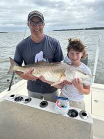 A proud father and son duo displaying a large Redfish on a fishing boat, showcasing their successful catch during a family fishing trip.