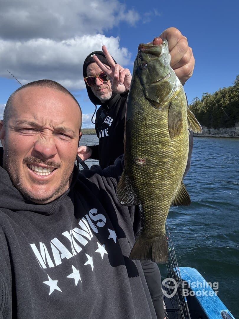 Two anglers proudly displaying a large Smallmouth Bass while fishing on a scenic lake.
