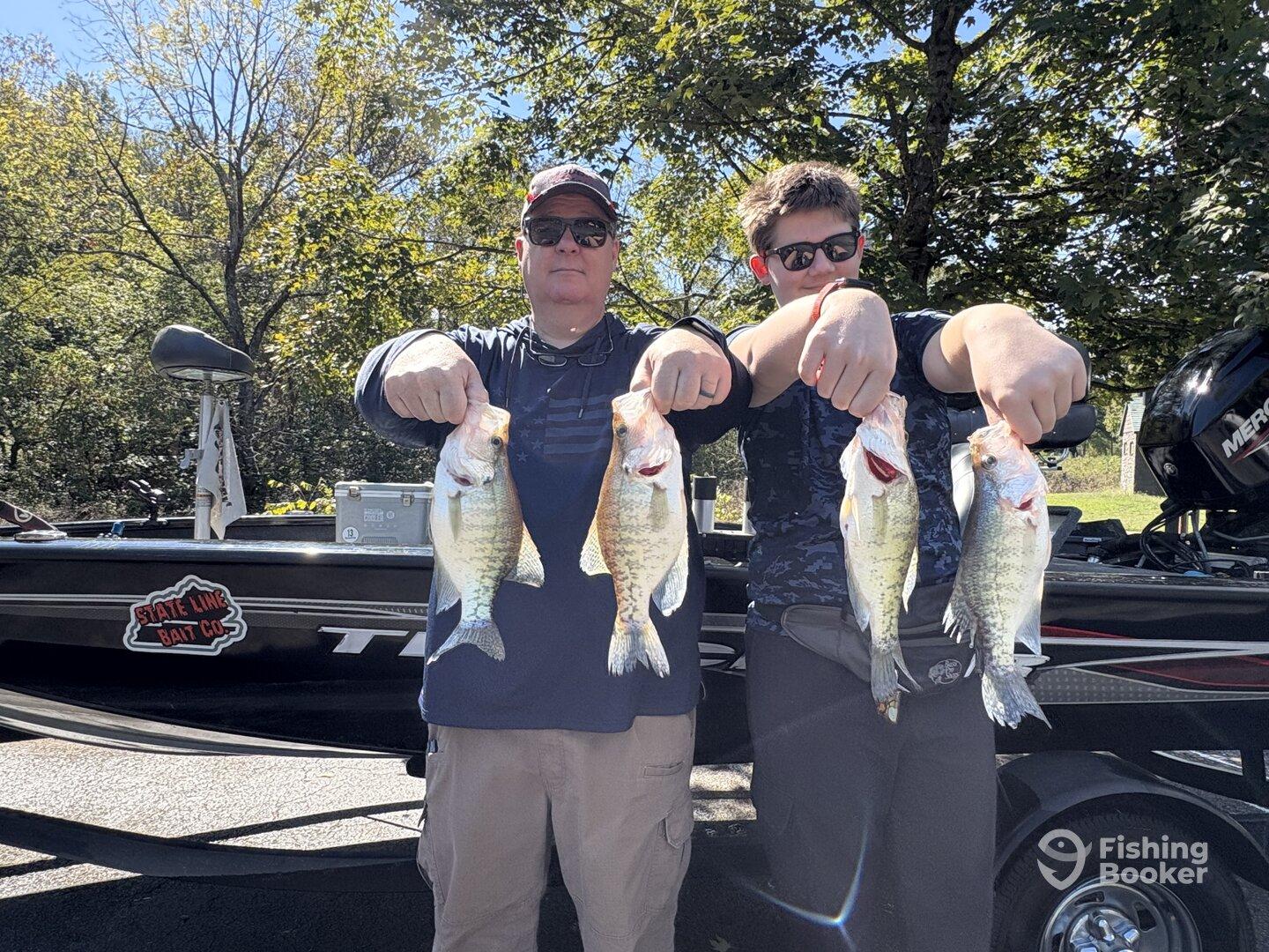 A proud adult and a young angler displaying their catch of several Largemouth Bass while standing by their fishing boat in a scenic outdoor setting.