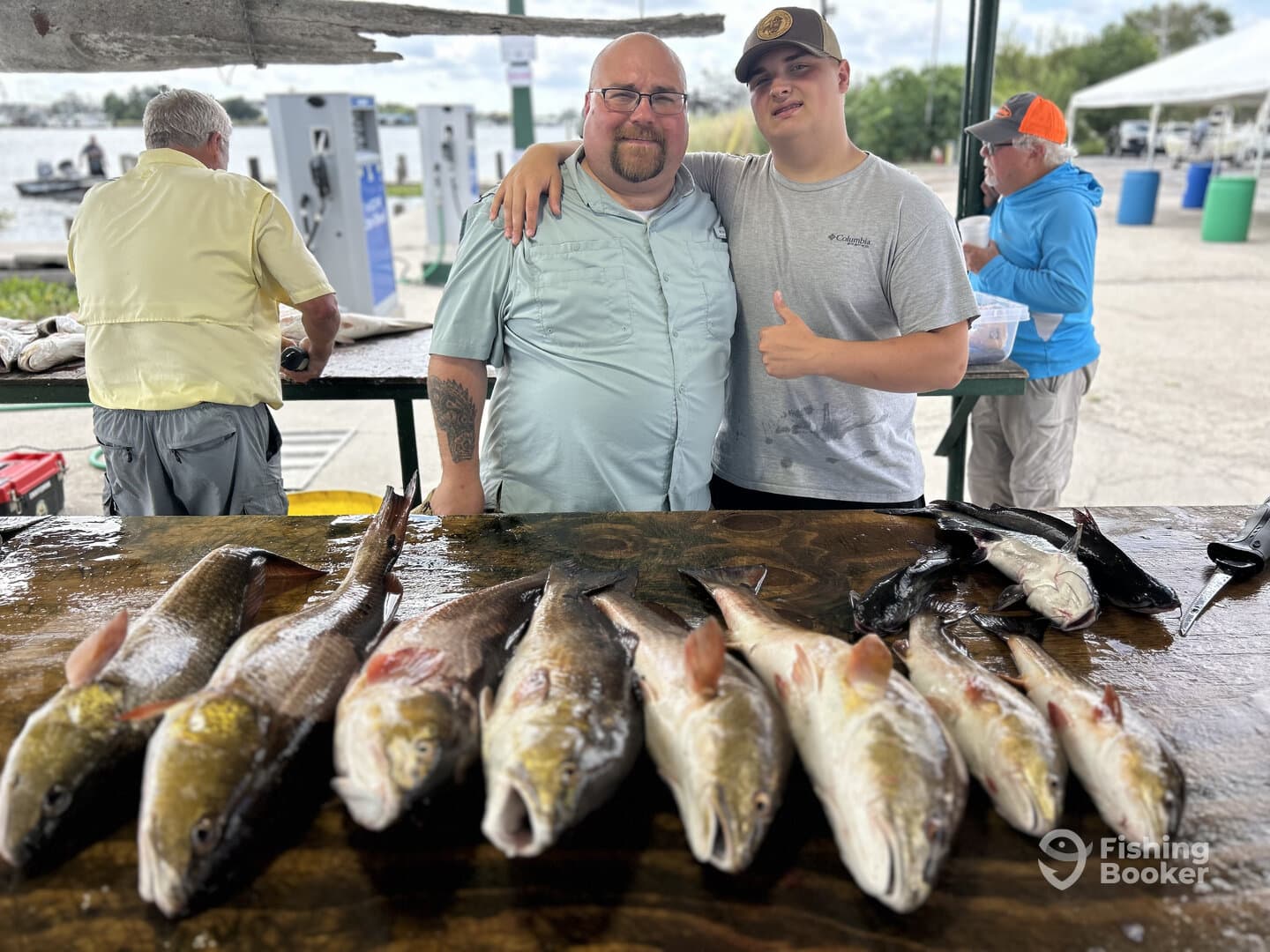 A proud duo poses with a variety of freshly caught fish on a display table at a fishing dock, showcasing their successful day on the water.