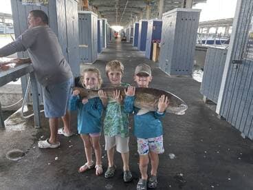 Three young anglers proudly displaying a large Redfish at a fishing dock, showcasing their successful catch after a fun day on the water.