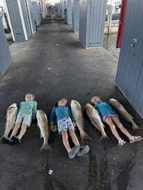 Two children lying next to several large Redfish on a dock, showcasing their catch after a successful fishing trip.